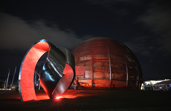 Nighttime view of the CERN Globe of Science and Innovation illuminated in red, with a sculpture in the foreground.