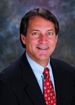 Headshot of Clayton Penhallegon, smiling in a dark suit, blue shirt, and red patterned tie against a mottled gray background.