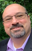Headshot of a bald man with glasses and a goatee, wearing a patterned button-up shirt, outdoors.