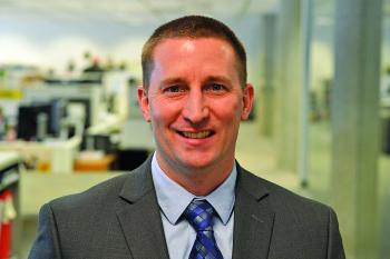 Luke Streit smiling in a gray suit and blue tie, photographed in front of a blurred office or lab background.